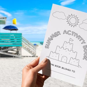 May include: A white summer activity book held in front of a beach scene. The cover features a line drawing of a sandcastle, sun, and clouds, with the words "Summer Activity Book". The book includes the text "This book belongs to:"