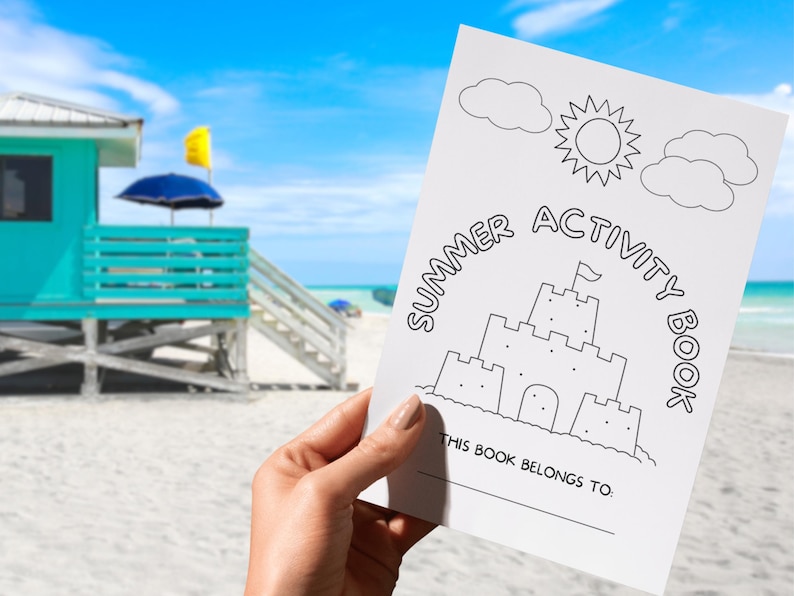 May include: A white summer activity book held in front of a beach scene. The cover features a line drawing of a sandcastle, sun, and clouds, with the words "Summer Activity Book". The book includes the text "This book belongs to:"