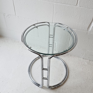 Peut inclure: Une table d'appoint circulaire dotée d'un plateau en verre transparent soutenu par une structure en métal chromé poli. La base se compose de deux tubes chromés circulaires entrelacés reliés par des barres métalliques verticales.
