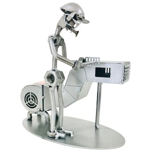 Puede incluir: Escultura metálica artesanal de un trabajador, construida con tuercas, pernos y otros elementos metálicos. La figura se representa en una postura de trabajo, con casco y herramientas, y está montada sobre una base ovalada. La escultura tiene un acabado plateado.