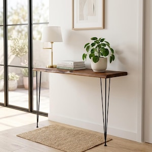 May include: A dark wood console table with black hairpin legs. A white lamp with a gold base, books, and a potted plant sit on the table. A woven rug is on the floor in front of the table. A framed picture hangs on the wall.