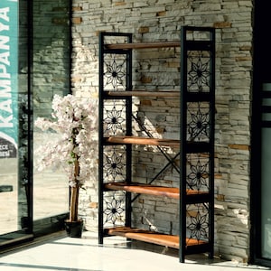 Puede incluir: Una estanter&iacute;a alta de metal negro y madera con seis estantes. El marco de metal negro presenta patrones decorativos de estrellas. Los estantes son de madera de color marr&oacute;n claro. Un &aacute;rbol de flores blancas est&aacute; a la izquierda de la estanter&iacute;a.