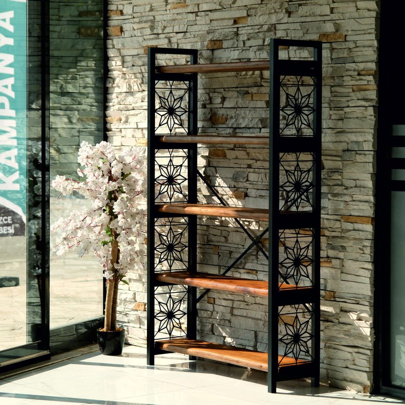 Puede incluir: Una estanter&iacute;a alta de metal negro y madera con seis estantes. El marco de metal negro presenta patrones decorativos de estrellas. Los estantes son de madera de color marr&oacute;n claro. Un &aacute;rbol de flores blancas est&aacute; a la izquierda de la estanter&iacute;a.