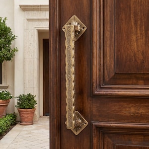 Può includere: Una maniglia per porta verticale in ottone con un intricato motivo floreale inciso e un bordo ondulato testurizzato. La maniglia è montata su una porta in legno marrone scuro con design a pannelli, dotata di piastre di montaggio quadrate.