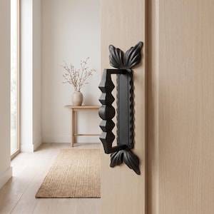 May include: A matte black metal door handle with a geometric, multi-faceted grip and a rectangular backplate featuring scalloped edges. The handle is mounted on a light-colored wooden door and terminates in decorative leaf-shaped end caps.