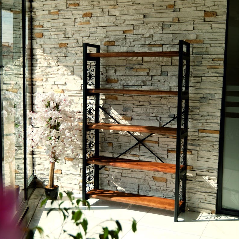 Puede incluir: Una estanter&iacute;a alta y abierta, con una estructura de metal negro y estantes de madera. Los estantes son de color marr&oacute;n c&aacute;lido, y el marco tiene recortes decorativos. La estanter&iacute;a est&aacute; contra una pared de piedra.