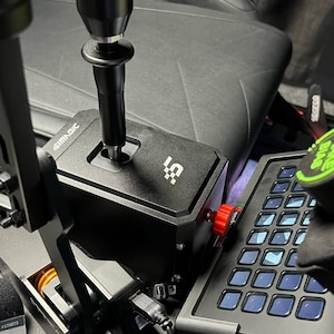 Puede incluir: Una palanca de cambios negra con una perilla roja y una base negra con el texto "SIMMAGIC" y un logotipo. La palanca de cambios está unida a un marco de metal negro. Un teclado negro con una cuadrícula de botones está en el fondo.