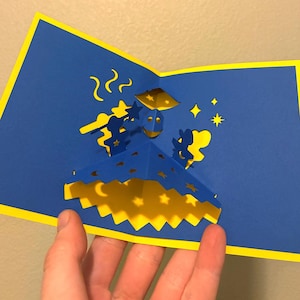 Op de afbeelding: Een pop-up wenskaart met een blauwe achtergrond en gele accenten. De kaart heeft een driedimensionaal ontwerp van een lachend gezicht met sterren en manen. De kaart is opengeklapt en onthult het ingewikkelde papierontwerp.