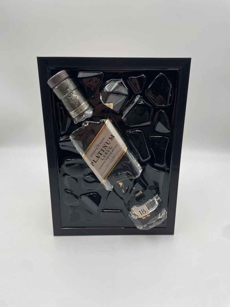Op de afbeelding: Een zwart omlijst kunstwerk met drie gedeeltelijk zichtbare whiskyflessen. De flessen zijn voorzien van de labels "Johnnie Walker Platinum Label" en "18". De achtergrond is een zwarte textuur.