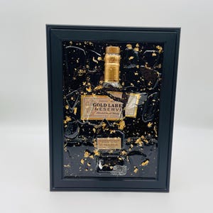 May include: A framed artwork featuring a Johnnie Walker Gold Label Reserve bottle. The bottle is set against a black background with gold leaf accents. The frame is black, and the artwork is displayed on a white background.