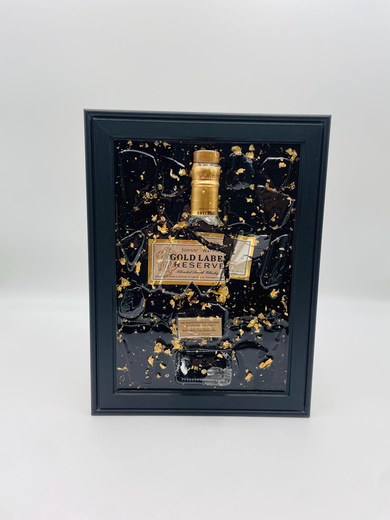 May include: A framed artwork featuring a Johnnie Walker Gold Label Reserve bottle. The bottle is set against a black background with gold leaf accents. The frame is black, and the artwork is displayed on a white background.