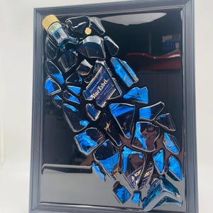 Op de afbeelding: Een ingelijst kunstwerk met een geconstrueerde Johnnie Walker Blue Label fles. Het stuk is samengesteld uit gefragmenteerd blauw glas en flesonderdelen, tegen een glanzende zwarte achtergrond in een zwart frame. Het etiket van de fles is zichtbaar.