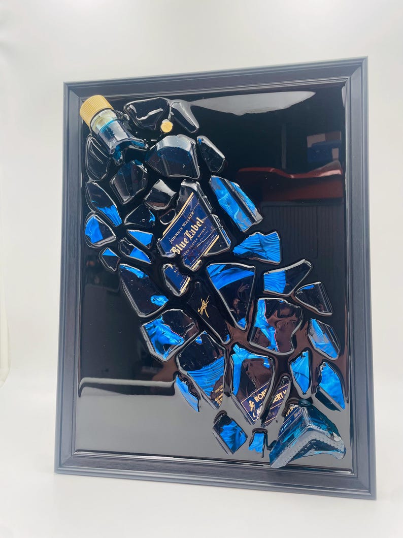 Op de afbeelding: Een ingelijst kunstwerk met een geconstrueerde Johnnie Walker Blue Label fles. Het stuk is samengesteld uit gefragmenteerd blauw glas en flesonderdelen, tegen een glanzende zwarte achtergrond in een zwart frame. Het etiket van de fles is zichtbaar.