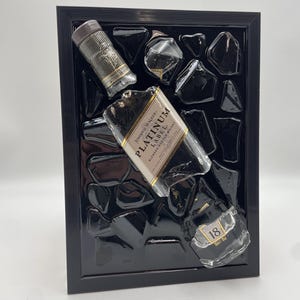 Op de afbeelding: Een zwart omlijst kunstwerk met fragmenten van Johnnie Walker Platinum Label blended scotch whisky flessen en zwarte hars. Het stuk bevat een dop en glasstukken, wat een textuurweergave cre&euml;ert.
