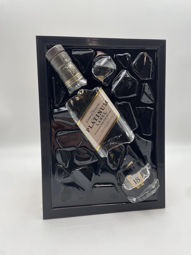 Op de afbeelding: Een zwart omlijst kunstwerk met fragmenten van Johnnie Walker Platinum Label blended scotch whisky flessen en zwarte hars. Het stuk bevat een dop en glasstukken, wat een textuurweergave cre&euml;ert.