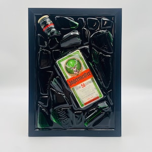 Pode incluir: Uma obra de arte emoldurada com um rótulo de garrafa Jägermeister e uma garrafa pequena, contra um fundo de estilhaços de vidro verde escuro. O rótulo é verde, branco e laranja, com o nome da marca e um emblema de veado. A moldura é preta.