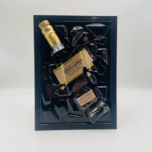 Pu&ograve; includere: Un'opera d'arte incorniciata con frammenti di una bottiglia di Johnnie Walker Gold Label Reserve. L'opera include il tappo, l'etichetta e pezzi di vetro rotti, il tutto su sfondo nero all'interno di una cornice nera.