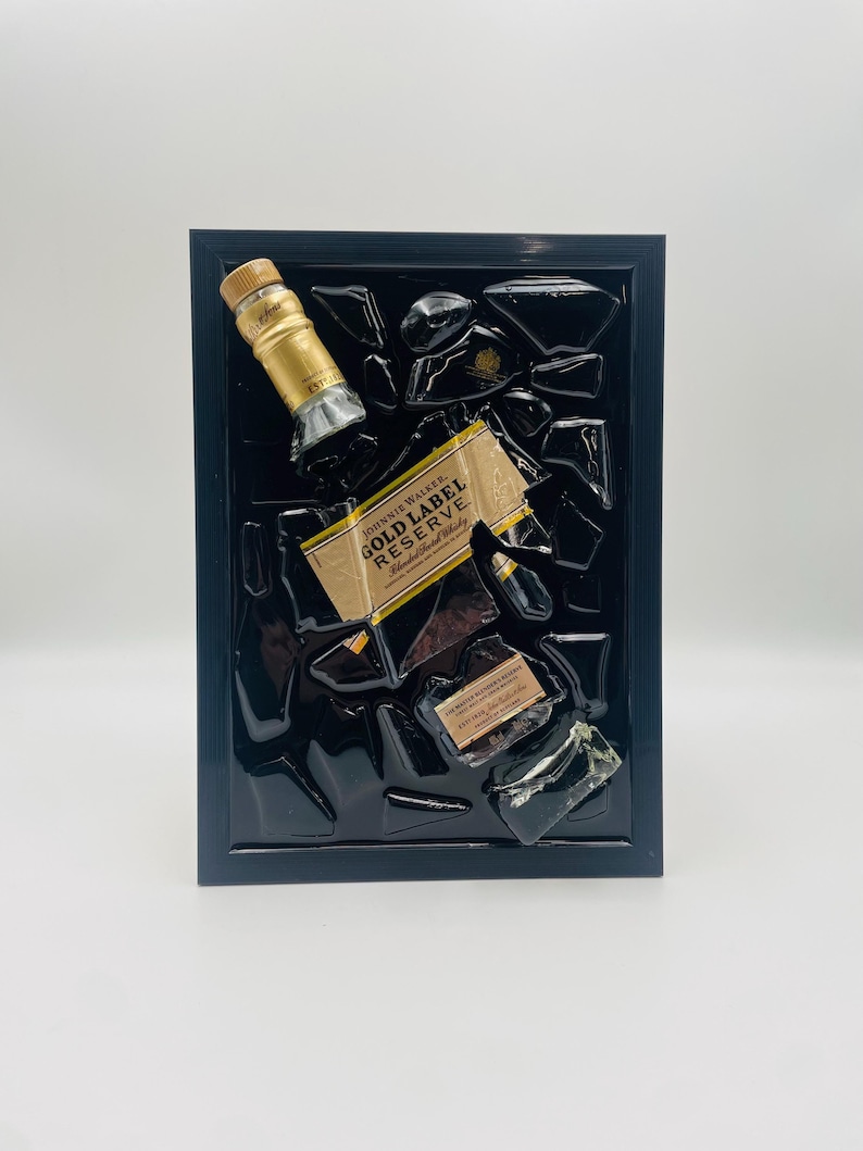 Pu&ograve; includere: Un'opera d'arte incorniciata con frammenti di una bottiglia di Johnnie Walker Gold Label Reserve. L'opera include il tappo, l'etichetta e pezzi di vetro rotti, il tutto su sfondo nero all'interno di una cornice nera.