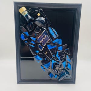 Op de afbeelding: Een ingelijst kunstwerk met een ontwerp van een gebroken fles. Het kunstwerk bevat fragmenten van een blauwe en zwarte fles, met het label "Blue Label" zichtbaar. De zwarte lijst versterkt de moderne, abstracte compositie.