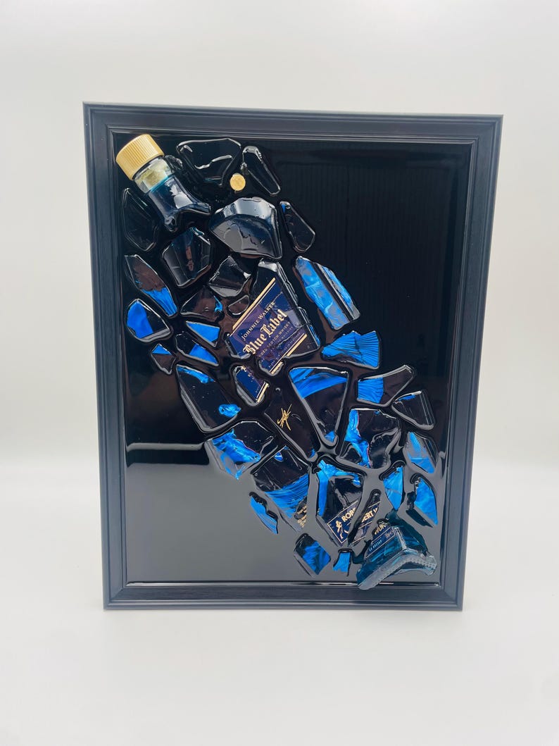 Op de afbeelding: Een ingelijst kunstwerk met een ontwerp van een gebroken fles. Het kunstwerk bevat fragmenten van een blauwe en zwarte fles, met het label "Blue Label" zichtbaar. De zwarte lijst versterkt de moderne, abstracte compositie.