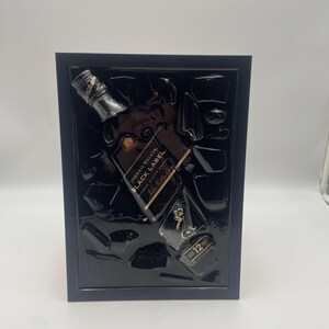 May include: A framed artwork featuring a black Johnnie Walker Black Label whiskey bottle and ice cubes. The bottle is angled, with gold lettering and a black label. The frame is dark blue, and the background is black.