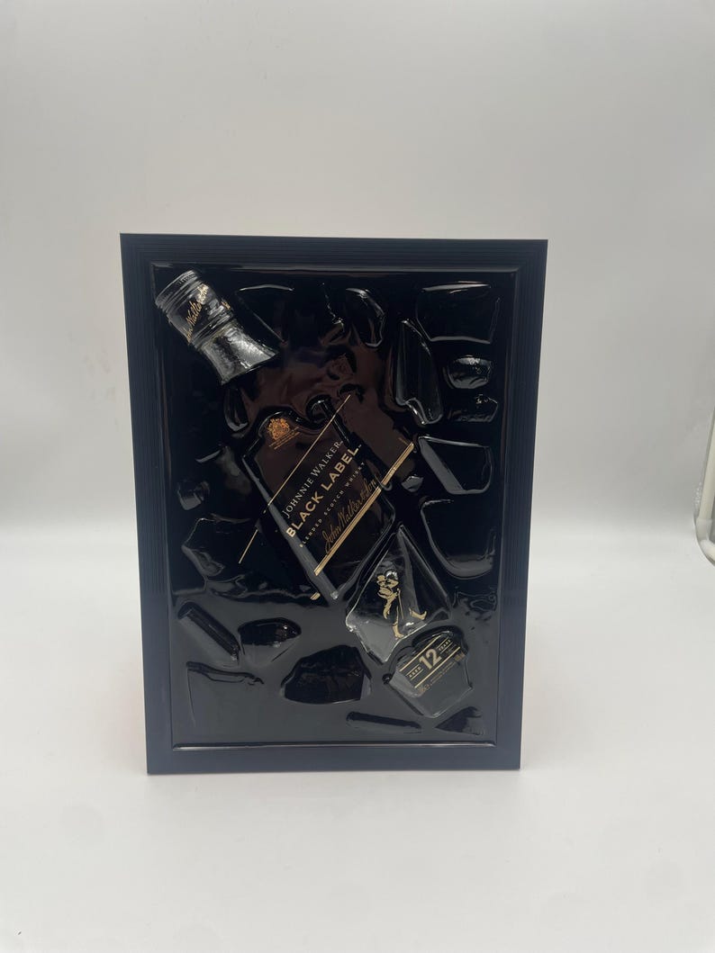 May include: A framed artwork featuring a black Johnnie Walker Black Label whiskey bottle and ice cubes. The bottle is angled, with gold lettering and a black label. The frame is dark blue, and the background is black.