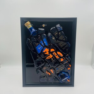 May include: A framed artwork featuring a collection of fragmented blue and black liquor bottles with orange accents. The artwork is encased in a black frame, creating a striking contrast against the white background. The bottles appear to be arranged in a dynamic, scattered composition.