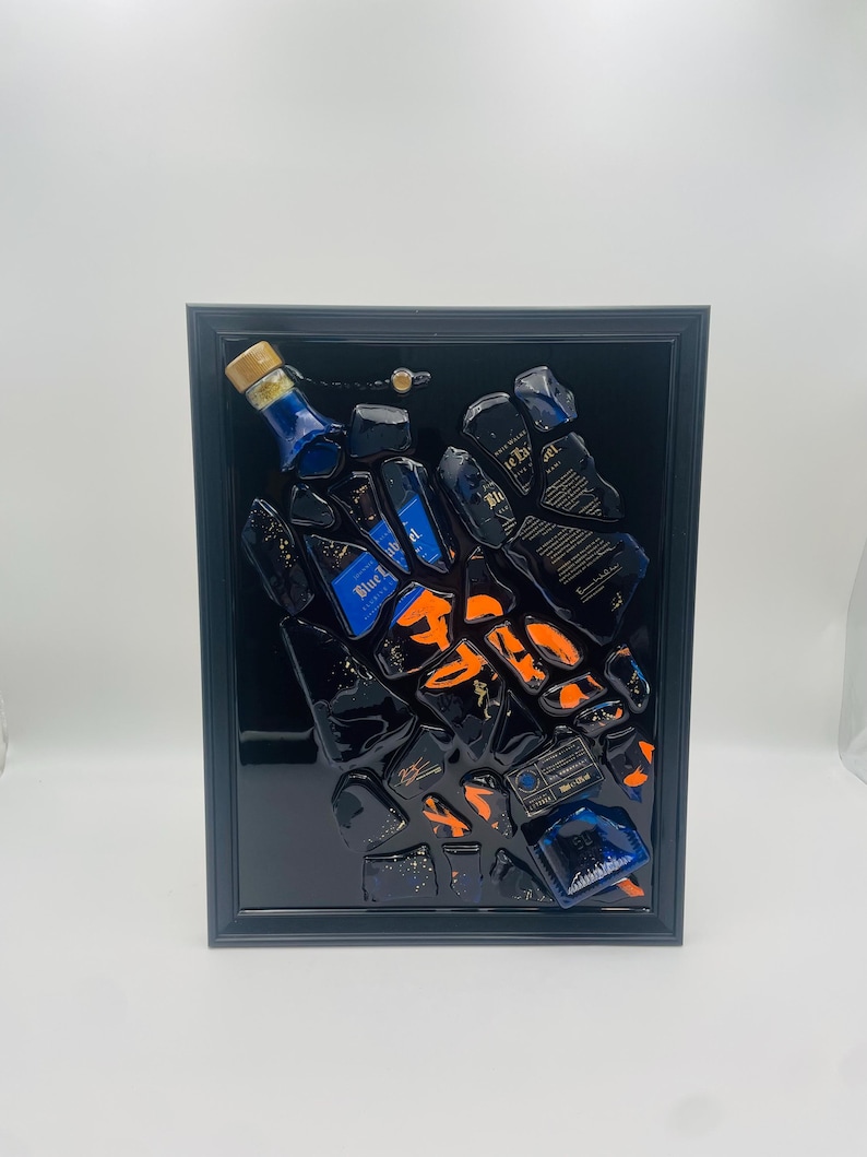 May include: A framed artwork featuring a collection of fragmented blue and black liquor bottles with orange accents. The artwork is encased in a black frame, creating a striking contrast against the white background. The bottles appear to be arranged in a dynamic, scattered composition.