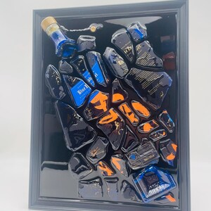 May include: Abstract artwork featuring fragments of a blue liquor bottle with orange accents, set against a black background within a framed display. The bottle's label is visible, adding detail to the composition. The artwork is a unique piece of home decor.