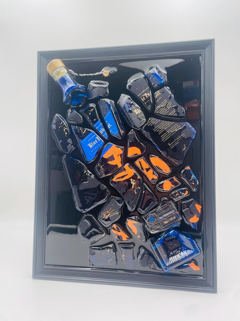May include: Abstract artwork featuring fragments of a blue liquor bottle with orange accents, set against a black background within a framed display. The bottle's label is visible, adding detail to the composition. The artwork is a unique piece of home decor.
