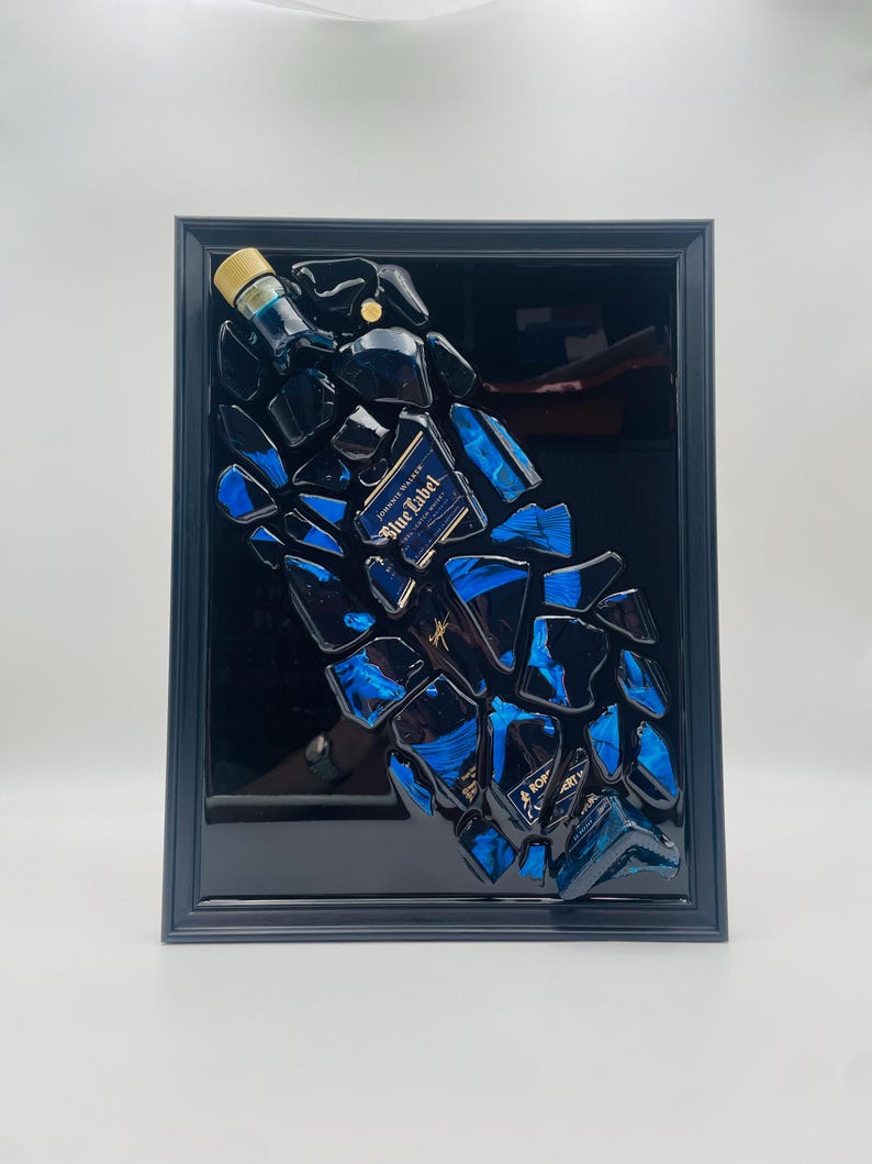 Op de afbeelding: Een ingelijst kunstwerk met een geconstrueerde fles Johnnie Walker Blue Label. Het kunstwerk bestaat uit gefragmenteerde stukken van de fles, met blauwe en zwarte tinten en het label "Blue Label". De lijst is zwart.
