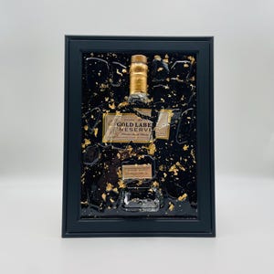 May include: Framed artwork featuring a Johnnie Walker Gold Label Reserve bottle. The bottle is encased in a black resin with gold leaf accents. The frame is black and rectangular, creating a striking contrast.