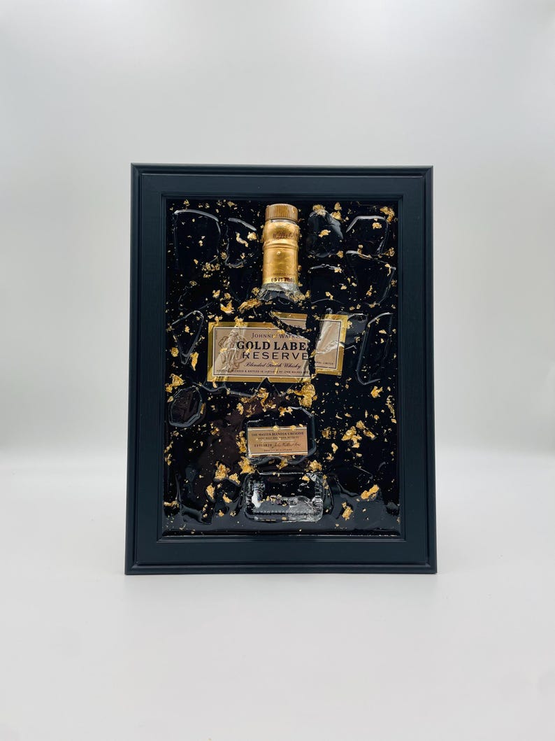 May include: Framed artwork featuring a Johnnie Walker Gold Label Reserve bottle. The bottle is encased in a black resin with gold leaf accents. The frame is black and rectangular, creating a striking contrast.