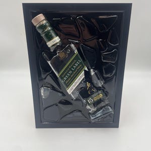 May include: A framed artwork with broken glass and three whisky bottles: Johnnie Walker Green Label and 15-year-old. The bottles are green and black, with gold labels. The frame is black.
