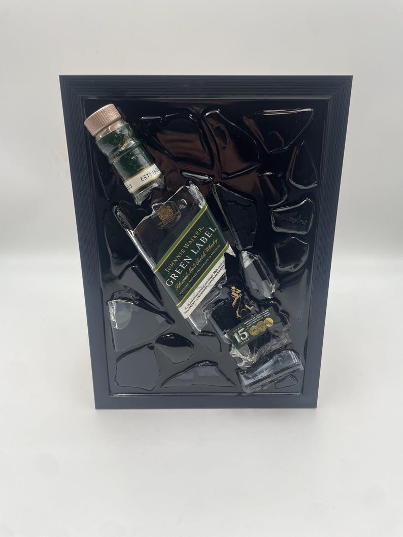 May include: A framed artwork with broken glass and three whisky bottles: Johnnie Walker Green Label and 15-year-old. The bottles are green and black, with gold labels. The frame is black.