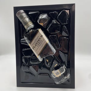 Op de afbeelding: Een zwart omlijst kunstwerk met een Johnnie Walker Platinum Label fles en andere glasstukken. De fles staat schuin, met het etiket zichtbaar. De achtergrond is een glanzend zwart oppervlak met onregelmatige vormen, wat een moderne esthetiek cre&euml;ert.