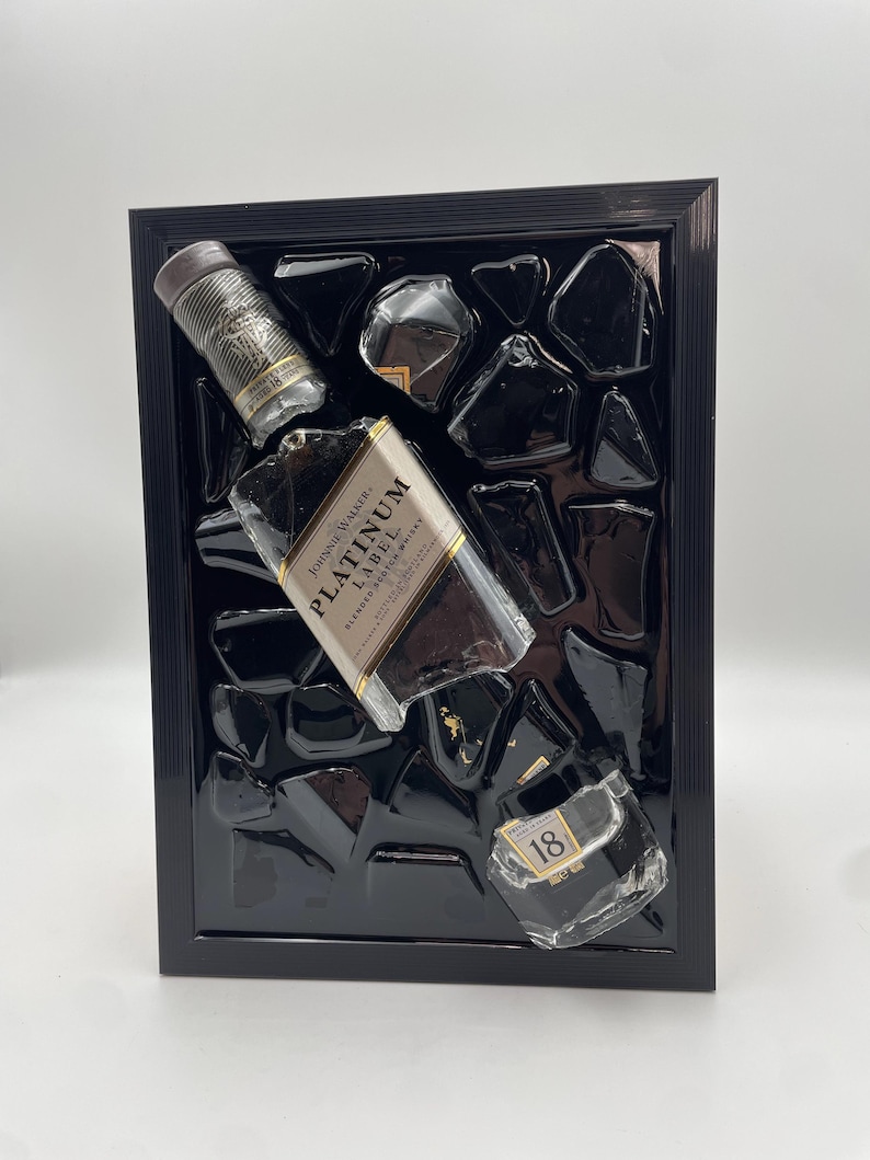 Op de afbeelding: Een zwart omlijst kunstwerk met een Johnnie Walker Platinum Label fles en andere glasstukken. De fles staat schuin, met het etiket zichtbaar. De achtergrond is een glanzend zwart oppervlak met onregelmatige vormen, wat een moderne esthetiek cre&euml;ert.