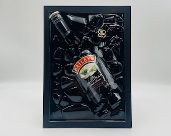 Op de afbeelding: Een ingelijst kunstwerk met een fles Bailey's Irish Cream likeur, een flesdop en decoratieve elementen. De fles is voorzien van het merk en de productbeschrijving. Het kunstwerk is geplaatst tegen een donkere achtergrond.