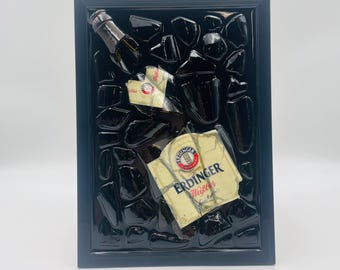 Op de afbeelding: Een ingelijst kunstwerk met een donkere achtergrond van glasscherven en stukken van een bierflesetiket. Het etiket luidt "ERDINGER Weißbier" in rode en witte tekst. Het frame is donkerblauw.