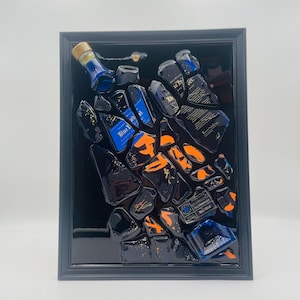 May include: Abstract art piece featuring fragments of blue and black liquor bottles with orange accents, set against a black background within a dark gray frame. The artwork showcases a unique, modern design.
