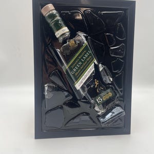 May include: A framed artwork depicting a broken Johnnie Walker Green Label whisky bottle. The piece includes fragments of the bottle and a small bottle, set against a black backdrop within a black frame, creating a dramatic effect.