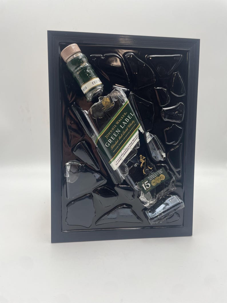 May include: A framed artwork depicting a broken Johnnie Walker Green Label whisky bottle. The piece includes fragments of the bottle and a small bottle, set against a black backdrop within a black frame, creating a dramatic effect.