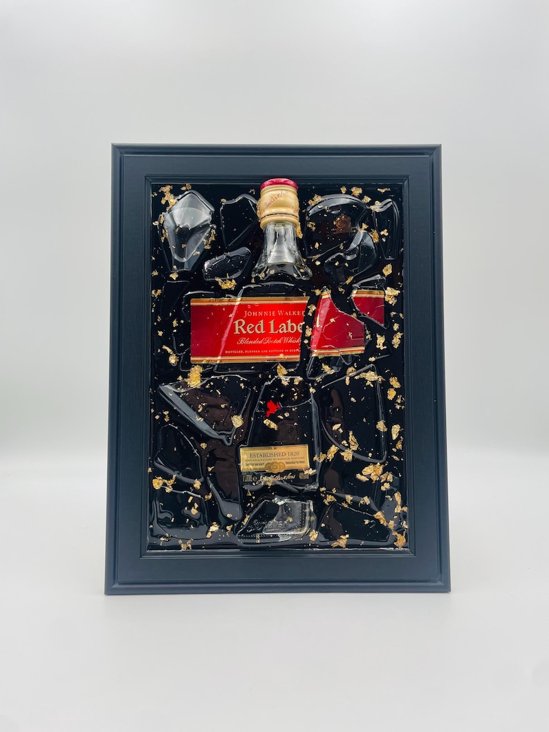 K&ouml;nnte beinhalten: Gerahmtes Kunstwerk mit einer Johnnie Walker Red Label Flasche. Die Flasche ist in schwarzem Harz mit Goldblatt-Akzenten und klaren Harzsplittern eingebettet. Der Rahmen ist dunkelblau.