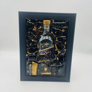 May include: Framed artwork featuring a Johnnie Walker Blue Label bottle. The bottle is centered against a black background with gold leaf accents, encased in a black frame. The label is visible, and the artwork has a textured appearance.