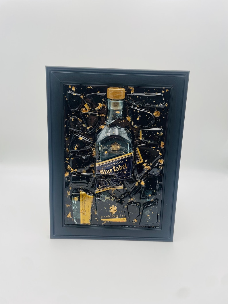 May include: Framed artwork featuring a Johnnie Walker Blue Label bottle. The bottle is centered against a black background with gold leaf accents, encased in a black frame. The label is visible, and the artwork has a textured appearance.