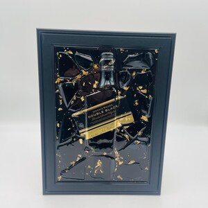 以下が含まれることがあります： ジョニーウォーカーダブルブラックのボトルをあしらった額装アート。黒いフレームは、金箔のアクセントと樹脂を施した黒い背景を囲んでいます。ボトルには金色のラベルが付いており、フレームの中央に配置されています。