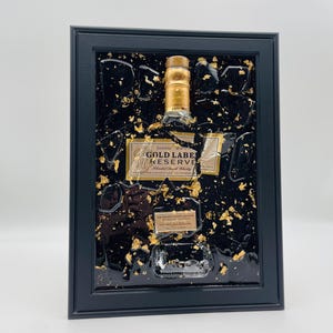 May include: A framed artwork featuring a bottle of Johnnie Walker Gold Label Reserve blended Scotch whisky. The bottle is set against a black background with gold leaf accents, encased in a black frame. The label is visible.