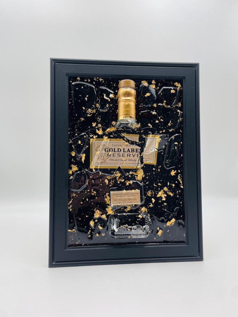 May include: A framed artwork featuring a bottle of Johnnie Walker Gold Label Reserve blended Scotch whisky. The bottle is set against a black background with gold leaf accents, encased in a black frame. The label is visible.