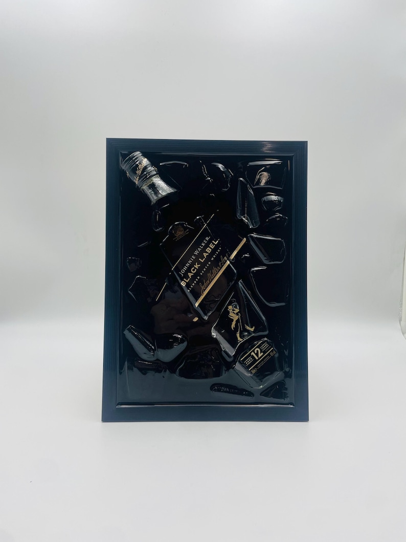May include: A framed artwork featuring a Johnnie Walker Black Label whiskey bottle and fragments. The black frame surrounds a dark background with the bottle and pieces of glass. The label is visible, and the number 12 is displayed.