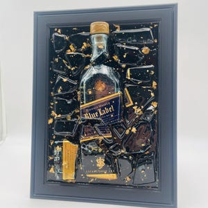 May include: Framed artwork featuring a Johnnie Walker Blue Label whiskey bottle. The bottle is set against a black background with gold leaf accents and encased in a dark blue frame. The label reads "Blue Label" and "Johnnie Walker".